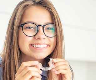 Teen with black glasses and long brown hair smiling holding Invisalign tray